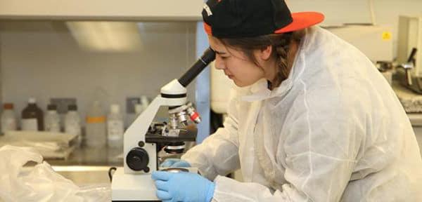 Young native woman looks into a microscope. She is wearing a protective cover over her clothes and hands.