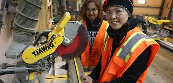 Two Alaska Native women wear bright orange vests and stand in front of a saw while measuring a board.