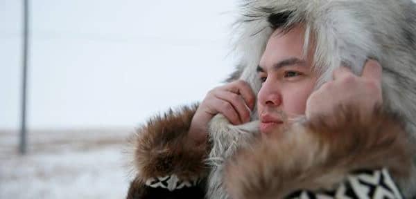 Young Alaska Native man holds up a parka hood around his face. He is outdoors.